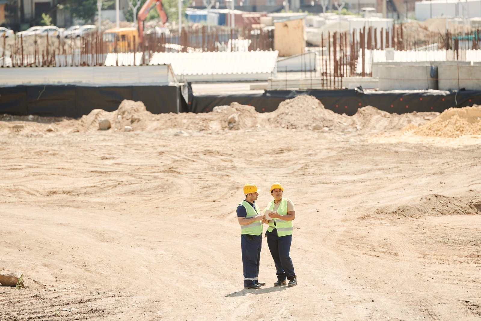 Two workers in high-visibility vests standing on a graded sand pad.