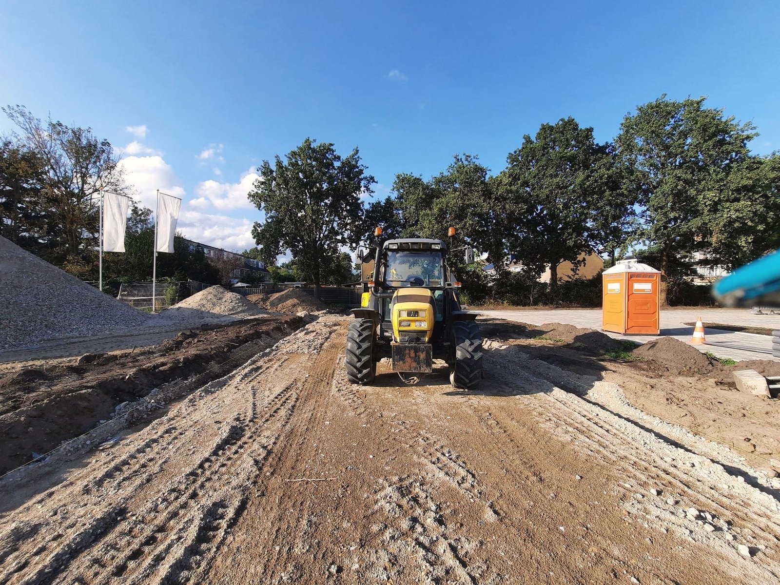 Tractor grading a dirt road with a portable restroom unit standing at the edge of the work.